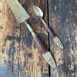 May include: A set of two silver-plated serving utensils with wooden handles. The utensils feature intricate carvings of a bird and a floral design. The utensils are on a wooden surface.