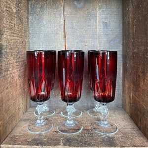 Peut inclure: Ensemble de quatre flûtes à champagne en verre rouge avec des tiges et des bases en verre clair. Les flûtes sont disposées en rangée sur une surface en bois.