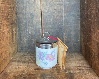 Puede incluir: Un pocillo de huevo de cerámica blanca con un diseño floral con flores rosas y moradas. El pocillo tiene una tapa plateada con un asa de anillo y una cuerda roja atada. El pocillo está sentado en una caja de madera con una etiqueta que dice "Coddled Eggs".