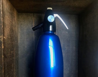 Pode incluir: Uma garrafa de vidro azul com uma bomba de cima preta e branca e uma alça branca. A garrafa está sentada em uma caixa de madeira.
