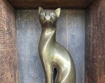 May include: A gold-colored metal cat figurine sitting in a wooden box. The cat is stylized and has a sleek, modern design.