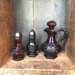 Peut inclure: Un ensemble de trois bouteilles en verre rouge rubis aux motifs complexes. Les bouteilles sont disposées sur une étagère en bois. La plus grande bouteille est un pichet avec une anse. Les deux plus petites bouteilles sont des salières et poivrières.
