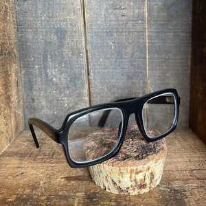 May include: Black plastic framed glasses with rectangular lenses, sitting on a piece of cork.