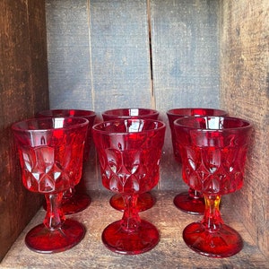 May include: Six red glass goblets with a diamond pattern are arranged in a row on a wooden surface. The goblets are all the same size and shape, and they have a clear, sparkling appearance.