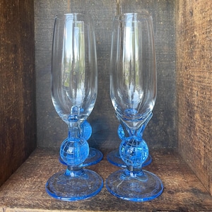 Può includere: Due set di flute da champagne in vetro blu con base a forma di globo. Le flute sono vuote e sono in una scatola di legno.