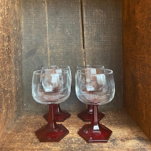 May include: Four clear wine glasses with ruby red hexagonal bases and stems. The glasses are displayed inside a wooden box. The glasses have a classic design, suitable for serving wine or other beverages. The glasses are made of clear glass.