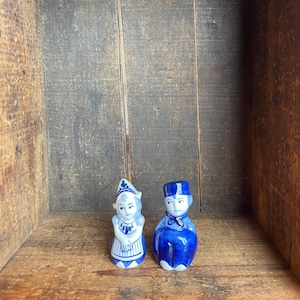 May include: Two ceramic figurines of a Dutch couple. The figurines are painted in blue and white, with the woman wearing a bonnet and apron, and the man wearing a hat and bow tie. The figurines are displayed in a wooden box.