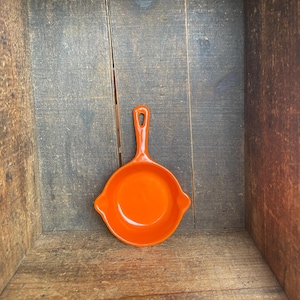 May include: A small, orange cast iron skillet with a single handle. The skillet is sitting in a wooden box.