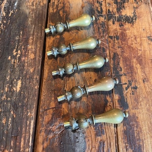 May include: Five antique brass drawer pulls with a wooden handle and decorative metal accents. The pulls are arranged on a weathered, brown wooden surface, showcasing their vintage design and craftsmanship. Each pull features a rounded handle and intricate metal detailing.