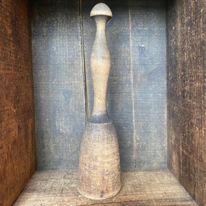 Puede incluir: Un mortero de madera vintage con una parte superior redondeada y un mango largo y texturizado. El mortero es de color marrón claro con áreas más oscuras, mostrando signos de desgaste. Se encuentra sobre un fondo de madera rústica.