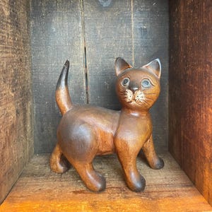 Könnte beinhalten: Eine Holzkatzenfigur mit glatter, polierter Oberfläche. Die Katze ist braun mit dunkleren Schattierungen auf Rücken und Schwanz. Sie hat detaillierte Gesichtszüge, einschließlich Augen und Schnurrhaare. Die Figur steht in einer Holzkiste.