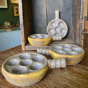 May include: Four ceramic escargot dishes with handles. The dishes have six circular indentations for the snails. The dishes are yellow and brown with a rustic, textured appearance. One dish is upright in a wooden box.
