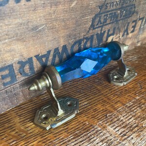 May include: A vintage door handle with a faceted, brilliant blue glass center. The handle has brass-colored metal hardware, including decorative mounting plates. The handle is set against a wooden surface with the text "STANLEY WORKS" visible.