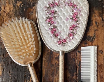 Puede incluir: Un conjunto de tres herramientas vintage para el cuidado del cabello: un espejo de mano blanco con un diseño floral, un cepillo de madera con cerdas naturales y un peine blanco.