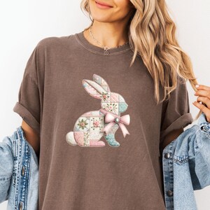 May include: Brown t-shirt featuring a patchwork bunny design with floral and pastel patterns. The bunny has a pink bow and is set against a neutral background. The shirt has rolled-up sleeves.