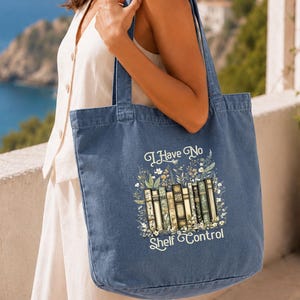 A woman is standing on a balcony, holding a blue tote bag with a printed design of books and the text "I Have No Shelf Control".