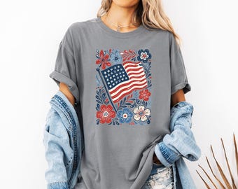 May include: A charcoal grey t-shirt featuring a central graphic of a waving American flag surrounded by stylized red, white, and blue floral and leaf illustrations. The design is printed on the front of the short-sleeved crew neck garment.