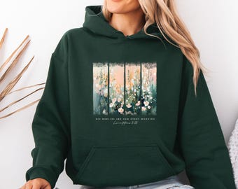 May include: A dark forest green hooded sweatshirt featuring a central graphic of abstract wildflowers in soft pink, white, and green tones. Below the floral design, the text reads: HIS MERCIES ARE NEW EVERY MORNING Lamentations 3:23.