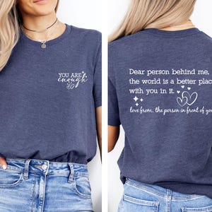 May include: A navy blue t-shirt with a white graphic on the front that says "YOU ARE enough" with a heart symbol. The back of the shirt has a white graphic that says "Dear person behind me, the world is a better place with you in it. love from, the person in front of you" with a heart symbol.