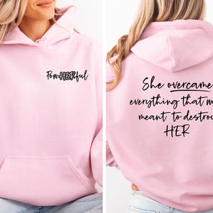 May include: A light pink hooded sweatshirt featuring a front pocket and black text on the chest that reads PowHERful. The back of the hoodie displays the black text She overcame everything that was meant to destroy HER in a cursive script font.