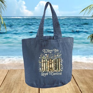A blue tote bag with a printed design of books and the text "I Have No Shelf Control" is displayed against a backdrop of a beach and palm trees.