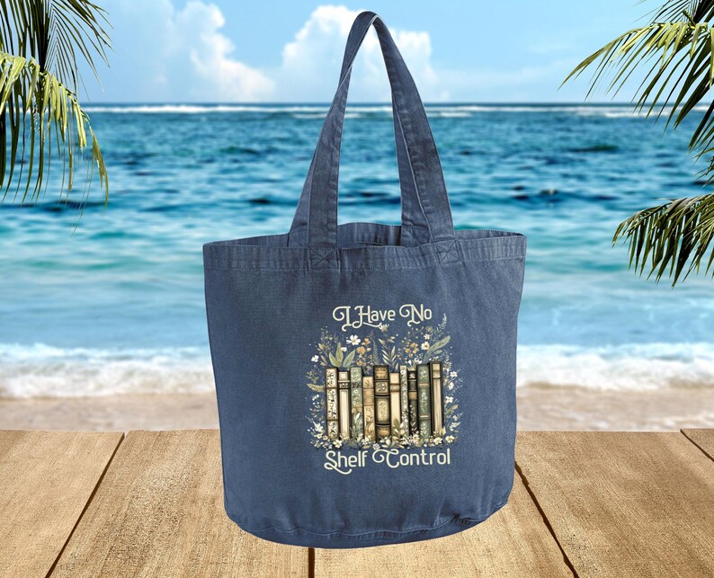 A blue tote bag with a printed design of books and the text "I Have No Shelf Control" is displayed against a backdrop of a beach and palm trees.