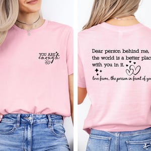 May include: Pink t-shirt with the text "YOU ARE enough" on the front and "Dear person behind me, the world is a better place with you in it. love from, the person in front of you" on the back.