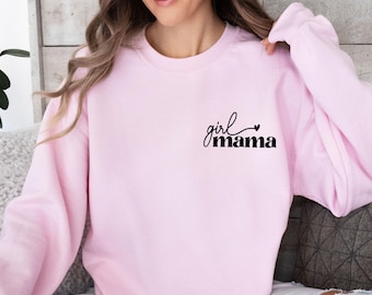 May include: A light pink long-sleeve crewneck sweatshirt featuring the black text "girl mama" printed on the upper left chest area. The script font for the word "girl" is positioned above the bold, sans-serif font for the word "mama".