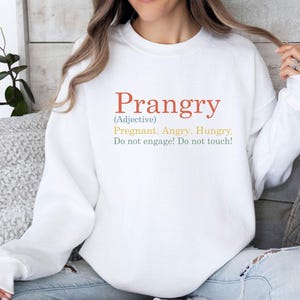 May include: A white crewneck sweatshirt featuring the text Prangry (Adjective) Pregnant. Angry. Hungry. Do not engage! Do not touch! The word Prangry is in orange, the definition words are in yellow, and the warning text is in green.