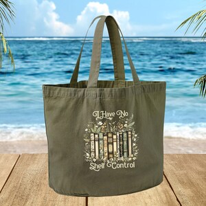 A green tote bag with a printed design of books and the text "I Have No Shelf Control" is displayed on a wooden surface, with a body of water and palm trees in the background.