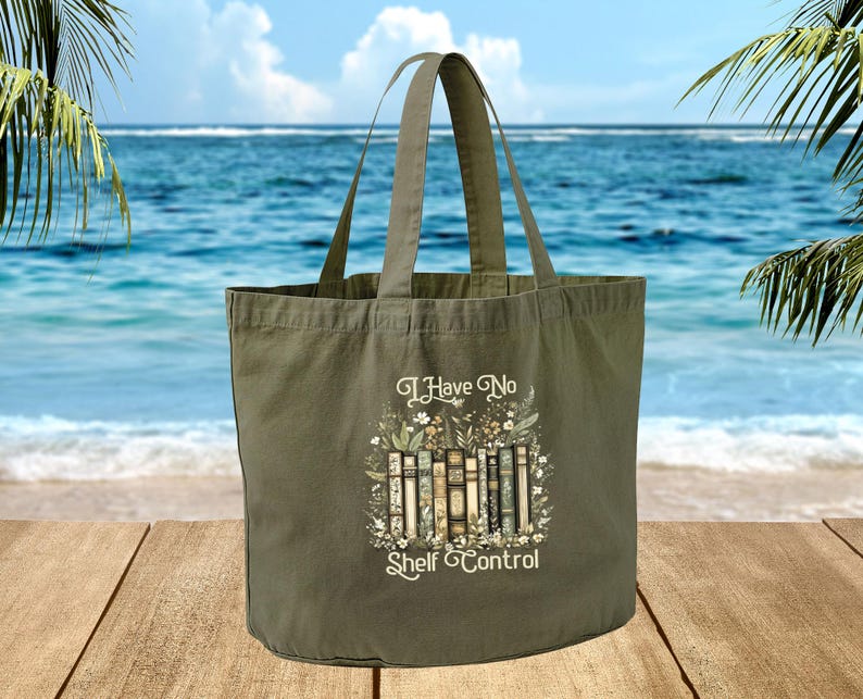A green tote bag with a printed design of books and the text "I Have No Shelf Control" is displayed on a wooden surface, with a body of water and palm trees in the background.