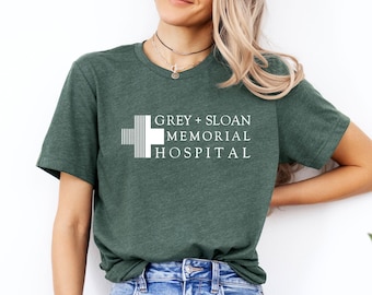 Puede incluir: Una camiseta de manga corta de color verde oscuro con un gráfico blanco de un símbolo de cruz y el texto GREY + SLOAN MEMORIAL HOSPITAL impreso en la parte delantera. La camiseta tiene cuello redondo y se combina con vaqueros azules.