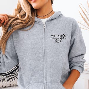 May include: A light gray zip-up hoodie with the text "YOU ARE enough" printed in black on the left chest. The hoodie has a drawstring hood and a full-length zipper. The model is wearing a white shirt underneath.
