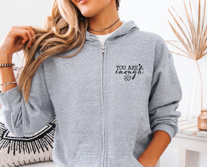 May include: A light gray zip-up hoodie with the text "YOU ARE enough" printed in black on the left chest. The hoodie has a drawstring hood and a full-length zipper. The model is wearing a white shirt underneath.