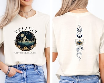 May include: A cream-colored t-shirt featuring a front graphic of a mountain inside a circle with the text VELARIS CITY OF STARLIGHT. The back of the shirt displays a vertical design of moon phases, floral elements, and a small mountain scene.