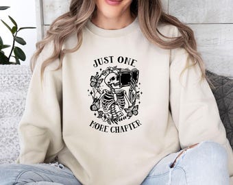 May include: A cream-colored crewneck sweatshirt featuring a black graphic print of a skeleton reading an open book, surrounded by butterflies, flowers, and stars. The text above and below the illustration reads, Just One More Chapter.