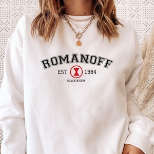 May include: A white long-sleeve crewneck sweatshirt featuring the black text ROMANOFF in an arched collegiate font, with EST 1984 flanking a red hourglass symbol inside a circle, and the words BLACK WIDOW printed in black below the symbol.
