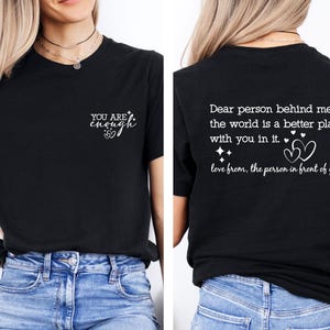 May include: Two black t-shirts with white text. The front shirt says "YOU ARE enough" with a small heart symbol. The back shirt says "Dear person behind me, the world is a better place with you in it. love from, the person in front of you" with two heart symbols.