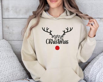 Puede incluir: Una sudadera con capucha de color beige que presenta un gráfico negro de astas de reno sobre el texto "merry Christmas" en letra cursiva, con un círculo rojo sólido situado debajo del texto que representa una nariz de reno.