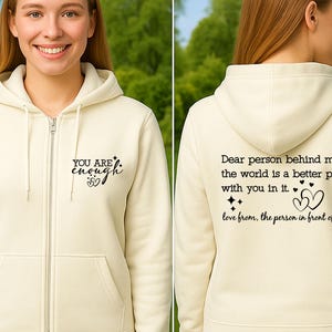 May include: Cream-colored zip-up hoodie with the text "YOU ARE enough" on the front. The back of the hoodie features the message "Dear person behind me, the world is a better place with you in it. love from, the person in front of you."