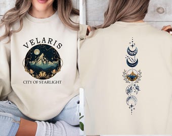 May include: A beige crewneck sweatshirt featuring a front graphic of a mountain inside a circle with the text VELARIS CITY OF STARLIGHT. The back displays a vertical design of moon phases, stars, and floral elements in blue and gold tones.