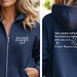 May include: Navy blue zip-up hoodie with white text that reads "YOU ARE enough" on the front. The back of the hoodie has the text "Dear person behind me, the world is a better place with you in it. love from, the person in front of you."