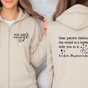 May include: A beige zip-up hoodie with black text on the front that reads "YOU ARE enough" with heart and star graphics. The back of the hoodie has the text "Dear person behind me, the world is a better place with you in it."