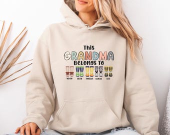 Puede incluir: Una sudadera con capucha de color beige que presenta el texto This GRANDMA BELONGS TO en letras coloridas sobre un gráfico de cinco pares de botas estampadas. Cada par de botas tiene los nombres Noah, Lilly, Amelia, Olivia y Leo debajo.