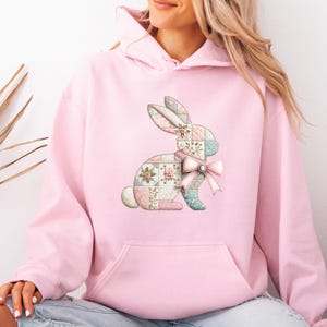 May include: A light pink hooded sweatshirt featuring a central graphic of a rabbit composed of a patchwork pattern in shades of pink, blue, and white. The rabbit design includes a light pink ribbon bow at the neck and floral embroidery details.