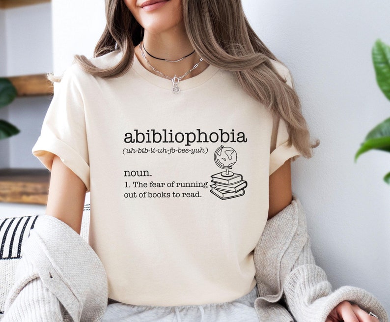 May include: A white t-shirt with a black graphic design that reads "abibliophobia (uh-bib-li-uh-fo-bee-yuh) noun. 1. The fear of running out of books to read."