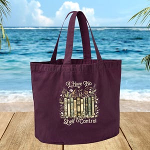 A maroon tote bag with a printed design of books and the text "I Have No Shelf Control" is displayed on a wooden surface, with a body of water and palm trees in the background.