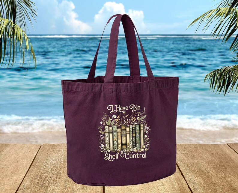 A maroon tote bag with a printed design of books and the text "I Have No Shelf Control" is displayed on a wooden surface, with a body of water and palm trees in the background.