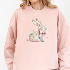 May include: A light pink crewneck sweatshirt featuring a central graphic of a rabbit composed of various pastel-colored patchwork squares with floral patterns. The rabbit is adorned with a light pink ribbon bow around its neck, tied in a knot.
