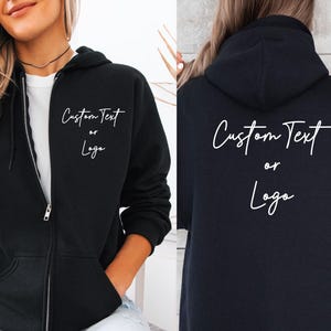 May include: Black zip-up hoodie featuring the text "Custom Text or Logo" in white script on the front and back. The hoodie has a hood, a full-length zip, and front pockets. The model is wearing a black choker.